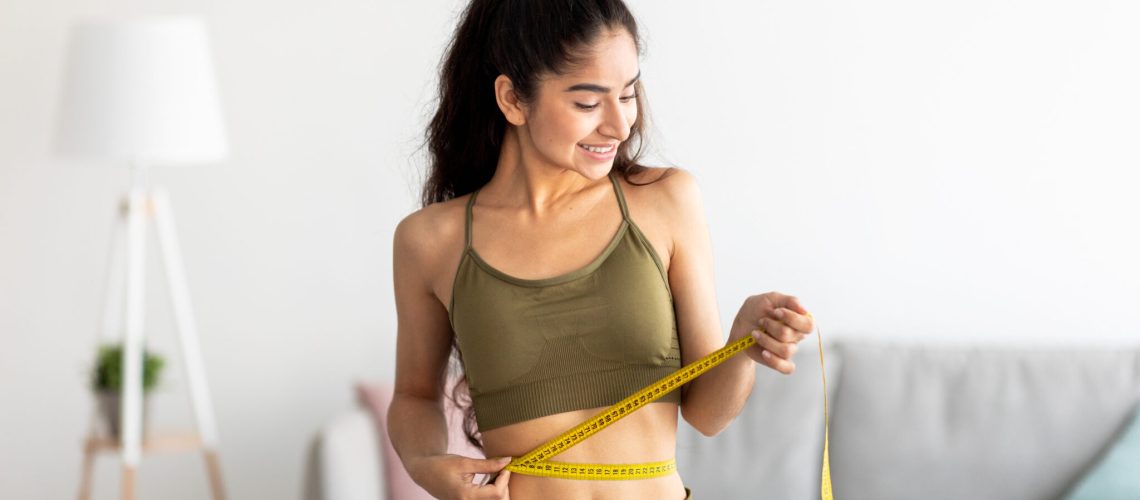 Successful weight loss concept. Fit young Indian woman in sports clothes measuring waist with tape at home. Millennial lady feeling satisfied with slimming program indoors